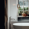 Freistehende Badewanne Spoon M von Agape in reinem Weiß aus Cristalplant biobased Mineralguss, Design Benedini Associati, in einem Badezimmer mit Fenster und bodenstehender Armatur in glänzendem Metall.