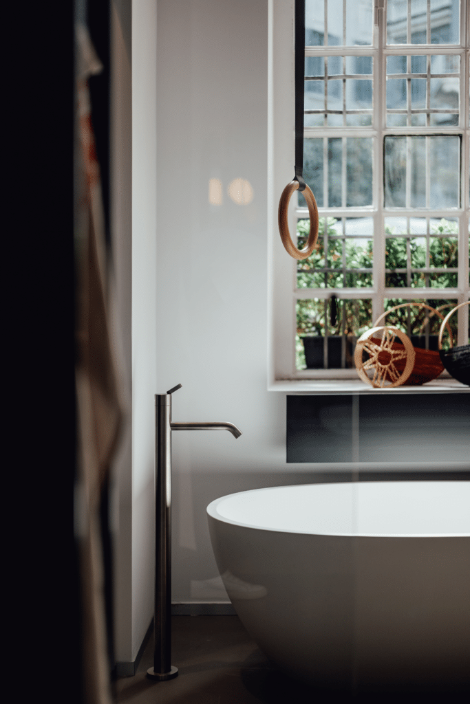 Freistehende Badewanne Spoon M von Agape in reinem Weiß aus Cristalplant biobased Mineralguss, Design Benedini Associati, in einem Badezimmer mit Fenster und bodenstehender Armatur in glänzendem Metall.